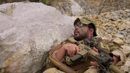 Fototapeta premium An exhausted soldier with face paint rests behind a rock, holding his rifle and catching his breath during a tactical military mission in a rugged environment.