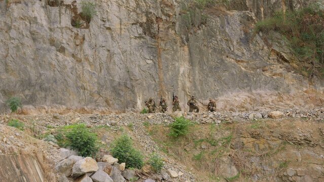 A military squad in camouflage takes a tactical position with rifles during a mission on a rugged cliffside, demonstrating teamwork and battlefield vigilance.