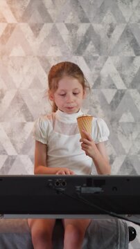 Close up of young girl eating ice cream while seated at electronic keyboard. Girl holds waffle cone with visible bite marks wearing white dress against geometric patterned wall backdrop