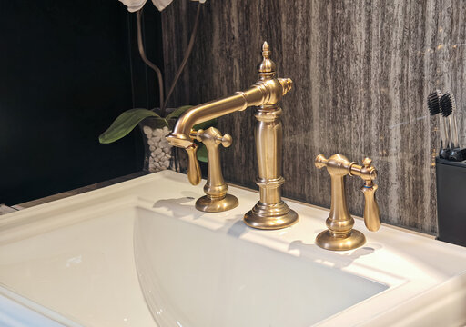 Luxury antique brass double handle mixer faucet installed on white ceramic washbasin with orchid flower and dark slate wall backdrop, targeted for luxury bathroom interior design