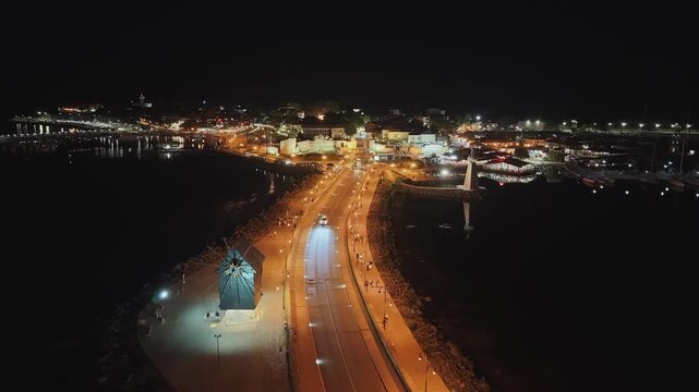 Night aerial 4K drone video of the narrow isthmus leading to the ancient town of Nesebar, Bulgaria. Features the historic wooden windmill, street lights, and sea view under the night sky.