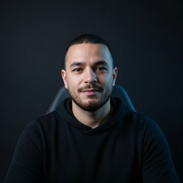 Retrato de primer plano de un joven profesional, programador o estudiante hispano con sudadera negra, mirando a la c&aacute;mara con iluminaci&oacute;n de estudio moderna sobre fondo oscuro.