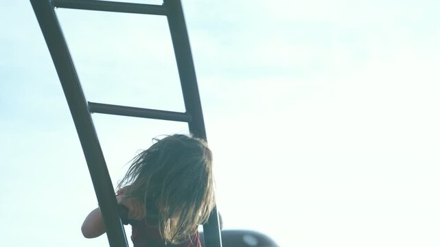 Rear view of a boy climbing a metal playground ladder silhouetted against bright sky