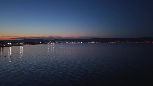 Twilight aerial 4K drone shot of the quiet Black Sea bay between Nesebar and Sunny Beach resort, Bulgaria. City lights reflecting on calm sea water during vibrant sunset horizon.