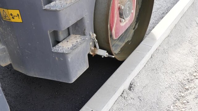 Road roller compacting newly paved asphalt layer adjacent to a concrete curb on an urban street construction and infrastructure site