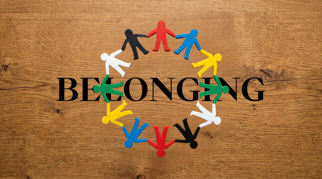 Colorful miniature figures holding hands in a circle surrounding the word "BELONGING" on a wooden background, representing diversity and team unity.
