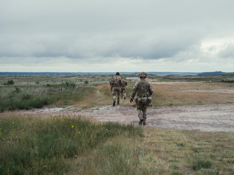 Three soldiers in British camouflage walk out across open heathland towards the firing point, helmets and daysacks under a heavy sky.