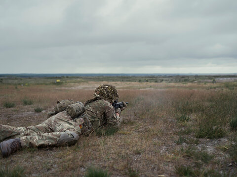 Wide view of a lone rifleman lying in rough grass on a military range, aiming downrange beneath a heavy overcast sky.