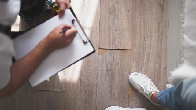 Top-down view of a professional repairman inspecting and measuring a damaged laminate floor with a tape measure, writing notes on a clipboard for a renovation estimate or report