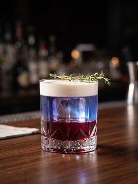 Layered Violet Craft Cocktail with White Foam Head and Thyme Garnish on a Dark Wooden Bar Counter