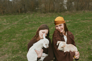Naklejka premium Two young women dressed in brown coats smile happily while holding white and light brown baby goats in a green meadow during a relaxed lifestyle moment outdoors.