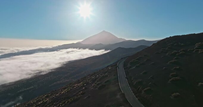 Mount Teide, Tenerife island, mountain peak rising above sweeping sea of clouds and fog during sunrise, with a winding road leading towards the volcano silhouette under a bright sun flare. Aerial shot