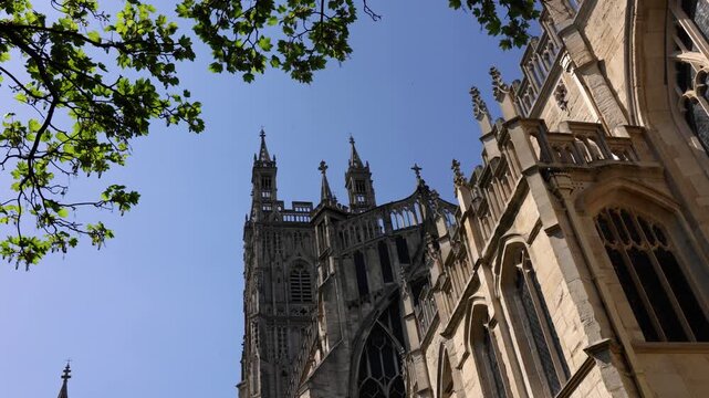 Horizontal 4K low angle shot of historic stone gothic towers and pinnacles framed by a bright green tree under a clear blue sky on a sunny day in the United Kingdom