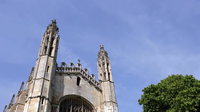 Horizontal 4K low angle shot of historic stone gothic towers and pinnacles framed by a bright green tree under a clear blue sky on a sunny day in the United Kingdom