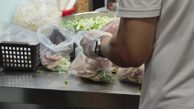Food vendor wearing gloves packing beef pho ingredients and meat into plastic bags for takeout at a local Vietnamese noodle shop
