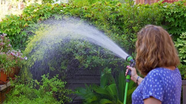 Slow motion woman watering dense garden foliage with a hose and wide fan of water