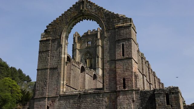 Horizontal 4K video of a majestic ruined stone abbey featuring a massive empty gothic arch framing a tower, with a bird flying under a clear blue sky