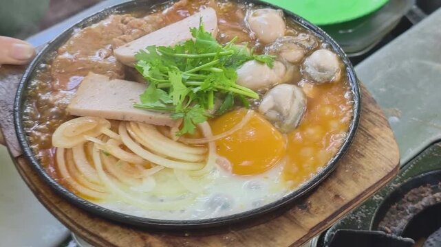Freshly cooked sizzling oyster dish in an iron pan being clamped and placed on a wooden tray to serve customers at a street food stall