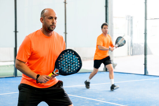 Men in orange shirts play doubles padel match indoors