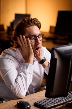 Young man in glasses shocked while looking at computer screen in dim office lighting, with man, young, glass, shocked detail, showing surprised, expression, mouthopen, handsonface context