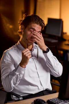 Stressed businessman rubbing eyes at desk in dim office after long workday, holding glasses with tired expression and warm blurred background during late evening