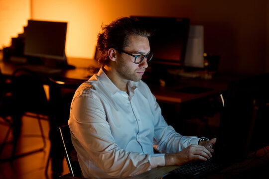 Stressed businessman rubbing eyes at desk in dim office after long workday, holding glasses with tired expression and warm blurred background during late evening