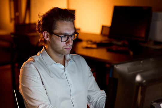 Stressed businessman rubbing eyes at desk in dim office after long workday, holding glasses with tired expression and warm blurred background during late evening