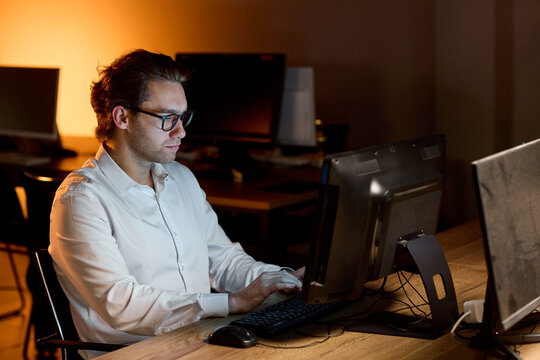 Stressed businessman rubbing eyes at desk in dim office after long workday, holding glasses with tired expression and warm blurred background during late evening