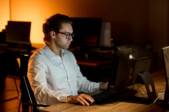 Stressed businessman rubbing eyes at desk in dim office after long workday, holding glasses with tired expression and warm blurred background during late evening