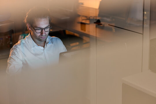 Middle aged man working on laptop in dim modern office seen through glass partition with warm lighting, reflections, and focused expression during late night work