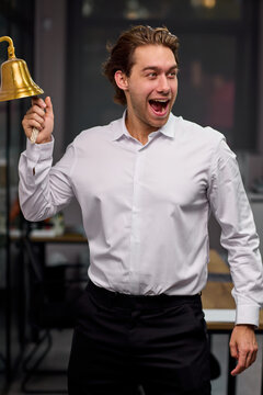 Young man ringing brass service bell with excited expression inside office or restaurant interior, with man, adult, male, shirt detail, showing dress, trouser, black, white context