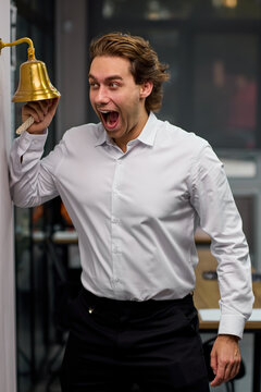 Young man ringing brass service bell with excited expression inside office or restaurant interior, with man, adult, male, shirt detail, showing dress, trouser, black, white context