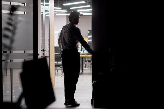 Silhouetted man opening glass door into brightly lit modern office interior at night, entering contemporary workplace with desks, chairs, overhead lighting and strong backlit contrast