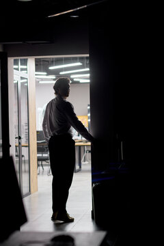 Silhouetted man opening glass door into brightly lit modern office interior at night, entering contemporary workplace with desks, chairs, overhead lighting and strong backlit contrast