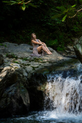 Fototapeta premium Young Woman Sitting on Mossy Rock Beside Waterfall in Lush Forest Stream, Relaxing Outdoors