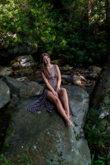 Fototapeta premium Portrait of Woman in Brown Dress Sitting on River Rock in Lush Forest Light
