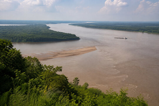 View from high bluff of wide brown river bending through low green forest, sandbar, tugboat and barge downstream, Mississippi River landscape