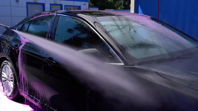 Professional car wash with pink foam and high pressure water spray on a black vehicle