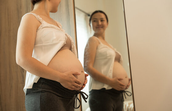 A pregnant woman in a white lace top smiles gently while holding her belly, her face clearly reflected in the mirror.