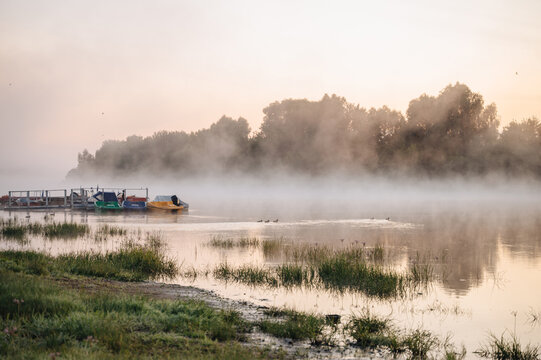 Misty sunrise over river with boats and summer reeds