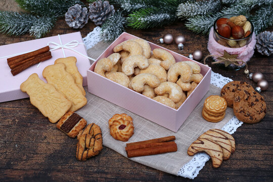  A pink gift box filled with traditional vanilla crescent cookies, surrounded by an assortment of Christmas cookies, cinnamon sticks, and decorations on a wooden table.
