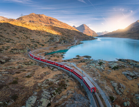 Aerial view of Bernina Express train near lake in the Swiss Alps at sunset, with glowing mountain peaks, water with reflections, rocky shore. Top drone view. Autumn landscape. Mountain train in fall