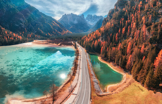 Aerial drone view of road along turquoise alpine lake in Dolomites, Italy. Coastal highway, mountain peaks, autumn larch trees forest slope, clear water, sky, clouds and reflection in fall. Top view