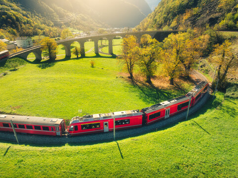 Aerial view of red train on Kreisviadukt in alpine mountains at sunset in autumn. Bernina Express, Switzerland. Top view of train, railroad, green grass, colorful trees in fall. Brusio spiral viaduct
