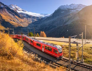 Red Bernina Express train passing through colorful autumn forest near glacier in the Swiss Alps, blue sky, golden trees and snowy mountain peaks. Travel in Switzerland. Red train and orange trees © den-belitsky