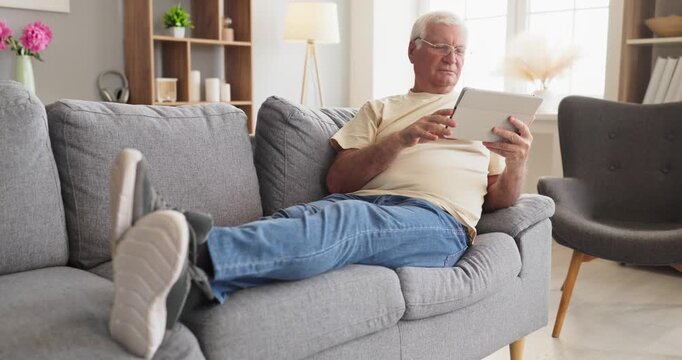 Senior man on sofa browsing tablet online. At home a relaxed retiree uses technology to read news chat and shop while reclining in a modern room. Clear concept of active aging in retirement.