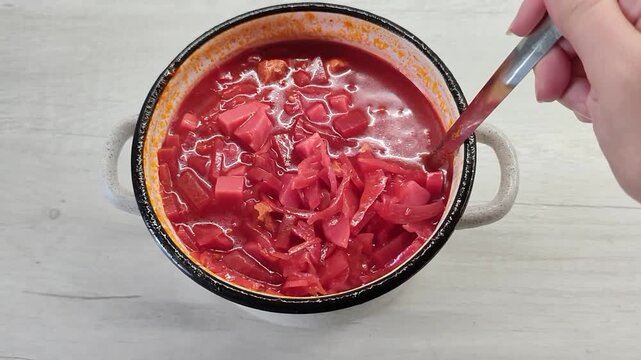Red borscht soup with cabbage and meat served in a small pot with a metal spoon on a light wooden table surface, top view