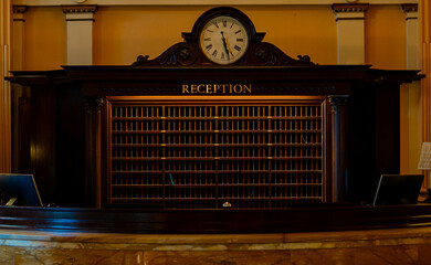 Hotel reception desk stands below vintage clock. Hotel reception area has dark wooden rack. Classic interior defines hotel reception. Vintage clock marks hotel reception desk. © Volodymyr