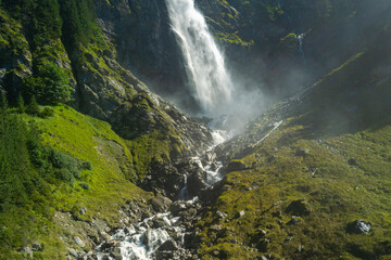Fototapeta premium Waterfall rushes through a deep alpine gorge. Waterfall cascades over steep mountain rocks. Rushing water surrounds waterfall below slope. Rocky landscape frames waterfall in summer.