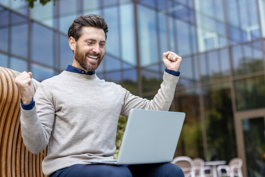 A happy young man sits on a bench, looks at the screen of a laptop he holds on his lap and rejoices with his hands raised up
